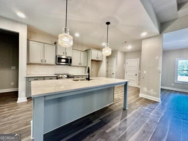 kitchen featuring dark wood finished floors, stainless steel appliances, decorative backsplash, an island with sink, and baseboards