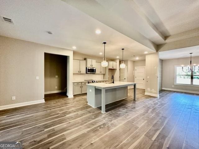 kitchen featuring wood finished floors, open floor plan, an island with sink, stainless steel microwave, and an inviting chandelier