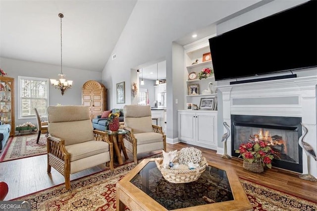 living area featuring built in features, a chandelier, a glass covered fireplace, wood finished floors, and high vaulted ceiling