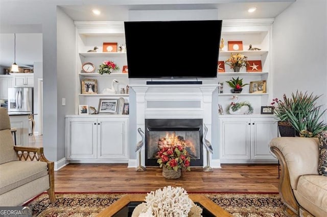 living area with a warm lit fireplace, built in shelves, wood finished floors, and recessed lighting