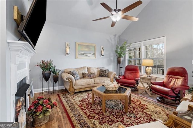 living room featuring a warm lit fireplace, high vaulted ceiling, ceiling fan, wood finished floors, and baseboards