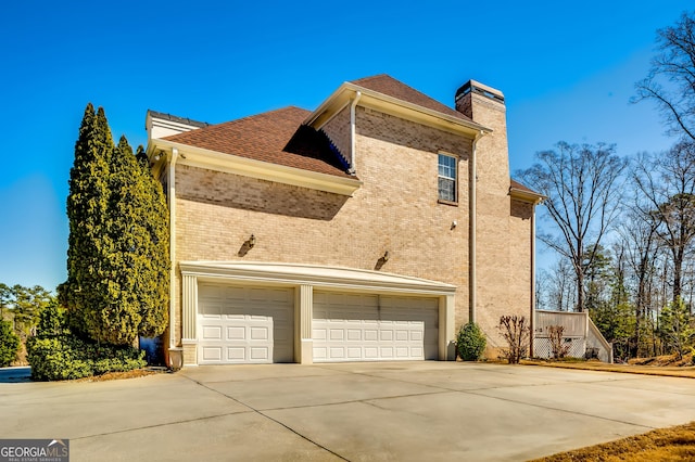 view of side of property with driveway, brick siding, a chimney, and an attached garage