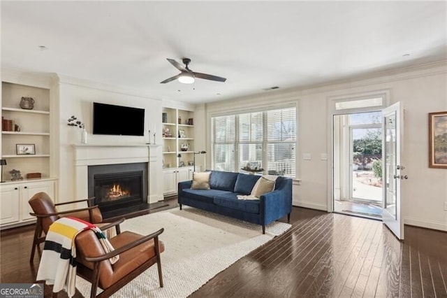 living room featuring built in shelves, a fireplace with flush hearth, baseboards, dark wood finished floors, and crown molding