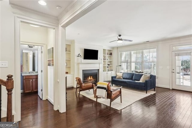 living area with dark wood-style floors, built in shelves, crown molding, a warm lit fireplace, and baseboards