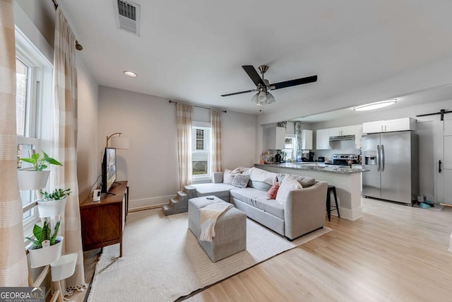 living area featuring visible vents, a barn door, a ceiling fan, light wood-style floors, and baseboards