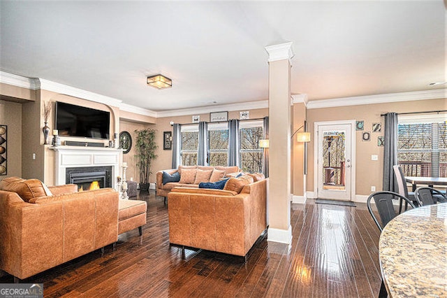 living area featuring a warm lit fireplace, dark wood-type flooring, baseboards, decorative columns, and crown molding