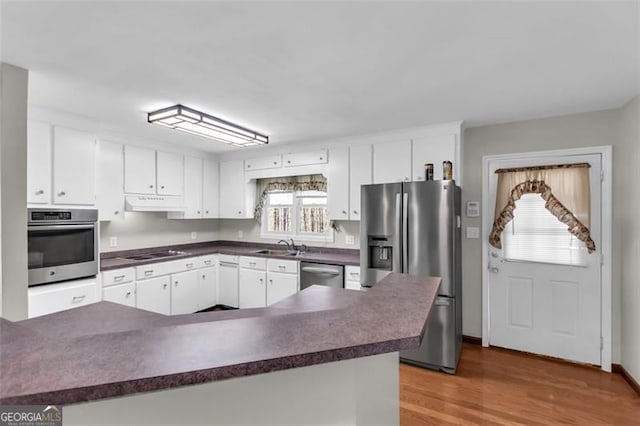 kitchen with under cabinet range hood, wood finished floors, white cabinetry, appliances with stainless steel finishes, and dark countertops