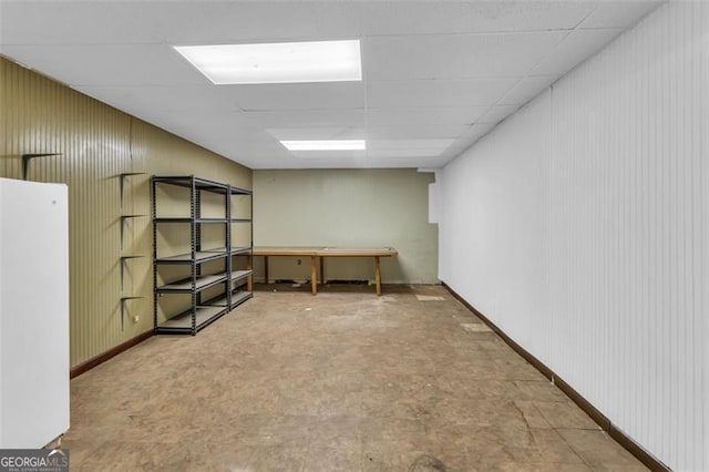 basement with a drop ceiling, freestanding refrigerator, and baseboards