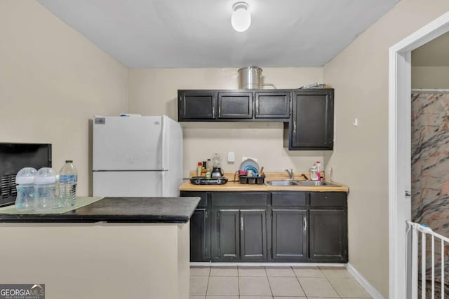 kitchen featuring dark countertops, dark cabinets, freestanding refrigerator, a sink, and light tile patterned flooring