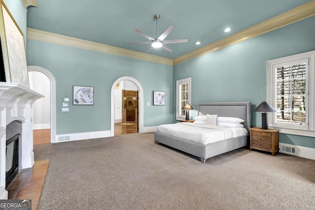 carpeted bedroom with visible vents, a fireplace with flush hearth, arched walkways, and crown molding