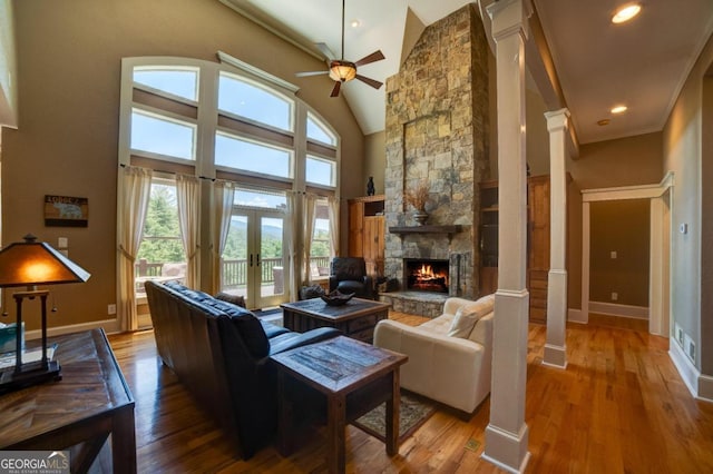 living room with baseboards, a towering ceiling, wood finished floors, a stone fireplace, and ornate columns