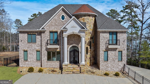 french provincial home featuring a balcony, brick siding, fence, french doors, and roof with shingles