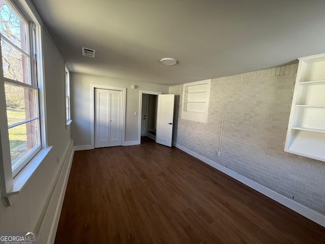 unfurnished bedroom featuring visible vents, brick wall, baseboards, a closet, and dark wood-style flooring