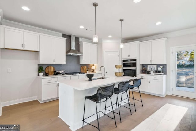 kitchen with multiple ovens, ornamental molding, white cabinets, a sink, and wall chimney range hood