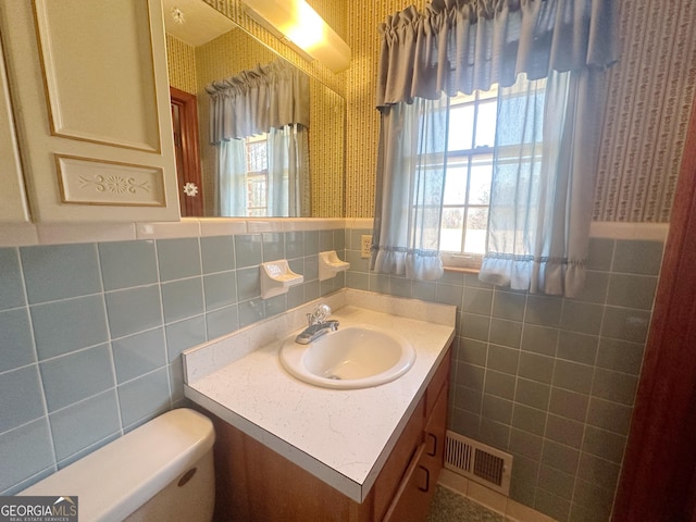 bathroom with wallpapered walls, visible vents, toilet, vanity, and tile walls