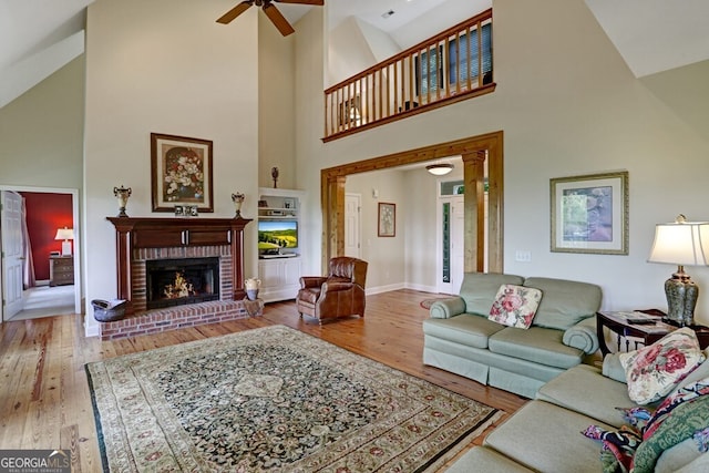 living room featuring baseboards, a towering ceiling, ceiling fan, hardwood / wood-style floors, and a brick fireplace