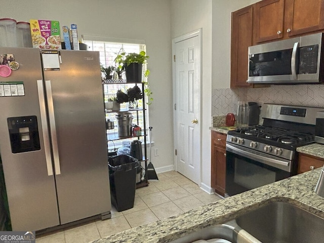 kitchen featuring light tile patterned floors, backsplash, appliances with stainless steel finishes, brown cabinetry, and light stone countertops