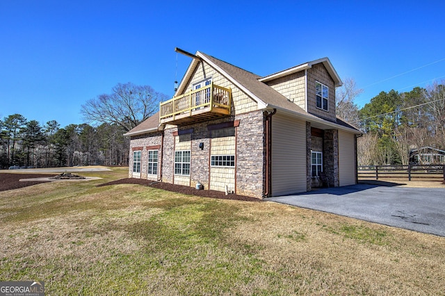 view of side of home featuring fence, aphalt driveway, a lawn, a balcony, and stone siding