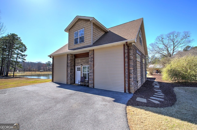 view of side of property featuring stone siding, aphalt driveway, a water view, and roof with shingles