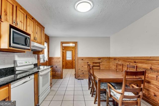 kitchen with white appliances, light tile patterned floors, a wainscoted wall, brown cabinets, and under cabinet range hood