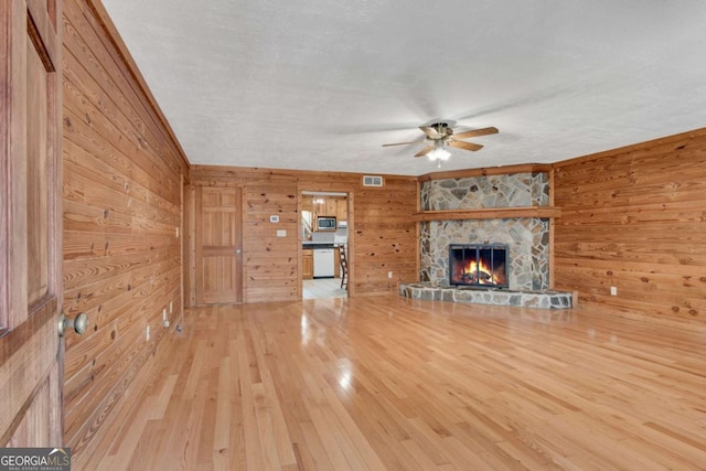 unfurnished living room featuring wooden walls, visible vents, ceiling fan, wood finished floors, and a stone fireplace