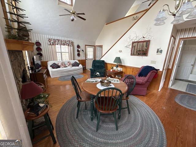 dining room with high vaulted ceiling, ceiling fan with notable chandelier, a wainscoted wall, and wood finished floors