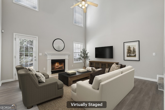 living area featuring baseboards, a ceiling fan, dark wood-style flooring, a lit fireplace, and a high ceiling