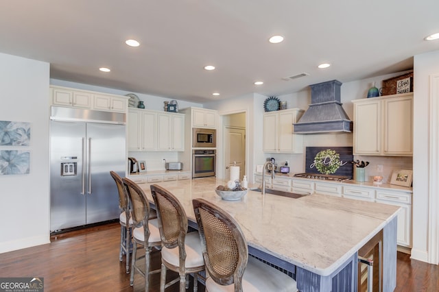 kitchen with a breakfast bar area, custom exhaust hood, a kitchen island with sink, a sink, and built in appliances