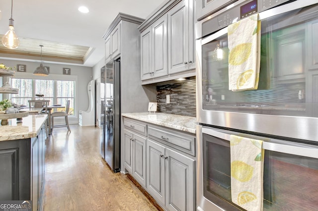 kitchen with a tray ceiling, gray cabinets, light wood-style flooring, decorative backsplash, and stainless steel double oven