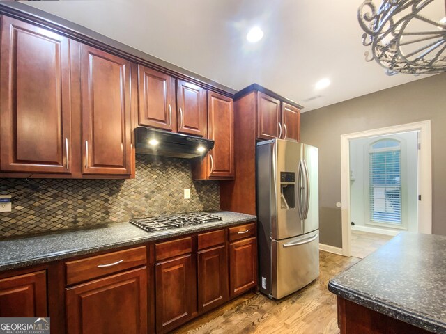 kitchen with light wood-style flooring, under cabinet range hood, appliances with stainless steel finishes, backsplash, and dark countertops