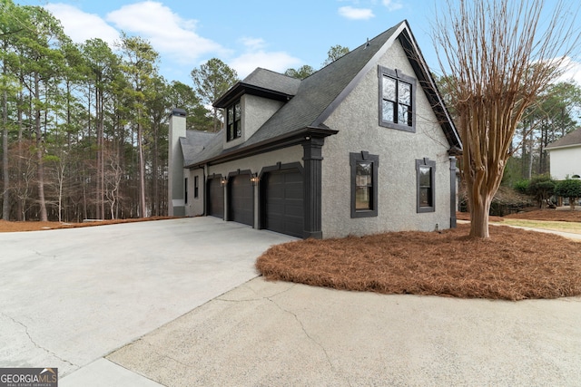 view of side of property featuring a garage, a shingled roof, concrete driveway, a chimney, and stucco siding