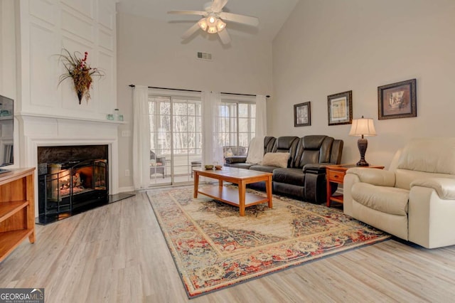 living area with a warm lit fireplace, visible vents, ceiling fan, wood finished floors, and high vaulted ceiling