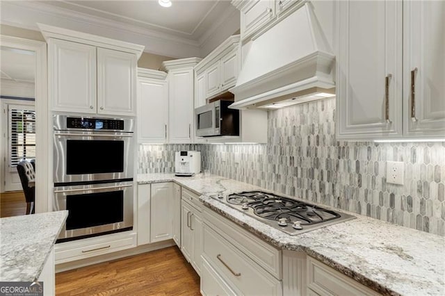 kitchen with white cabinetry, custom range hood, appliances with stainless steel finishes, light wood finished floors, and crown molding