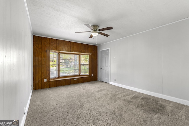 carpeted empty room featuring ceiling fan, a textured ceiling, ornamental molding, wooden walls, and baseboards