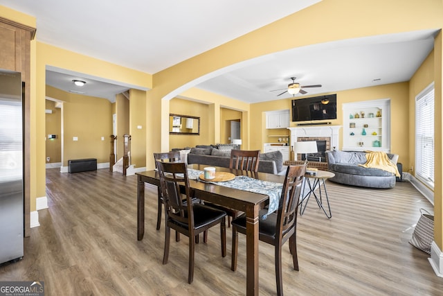 dining room with light wood-type flooring, ceiling fan, arched walkways, baseboards, and a brick fireplace