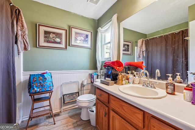 full bath with a textured ceiling, toilet, wood finished floors, visible vents, and wainscoting