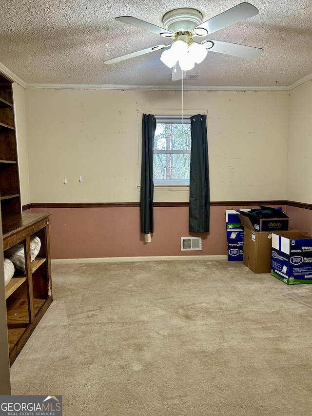 interior space with visible vents, a ceiling fan, a wainscoted wall, a textured ceiling, and crown molding