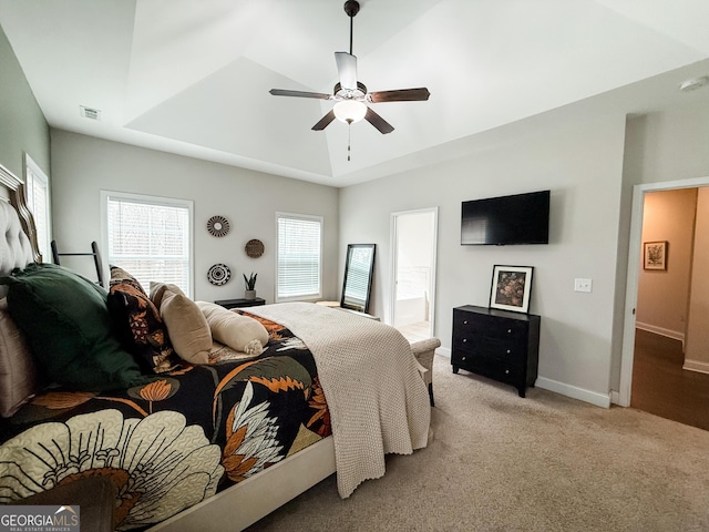 bedroom featuring baseboards, light colored carpet, ceiling fan, ensuite bathroom, and a tray ceiling