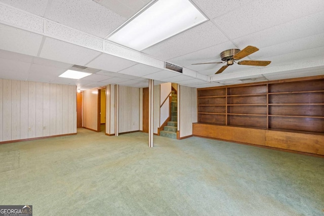 finished below grade area with built in shelves, carpet flooring, wood walls, ceiling fan, and stairs