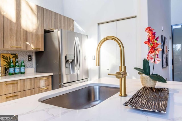 kitchen with tasteful backsplash, stainless steel fridge, brown cabinetry, light stone counters, and a sink