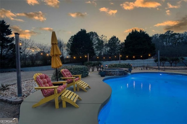 pool at dusk featuring a pool with connected hot tub, fence, and a patio