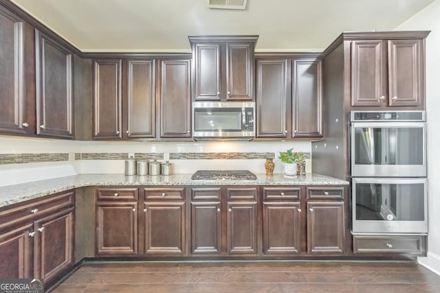 kitchen with light stone countertops, dark brown cabinetry, dark wood finished floors, decorative backsplash, and stainless steel appliances