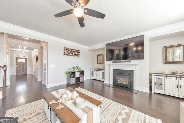 living room featuring dark wood-style floors, ceiling fan, wainscoting, a glass covered fireplace, and crown molding