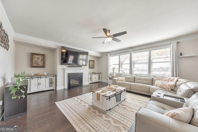 living room featuring ornamental molding, a glass covered fireplace, dark wood finished floors, baseboards, and ceiling fan