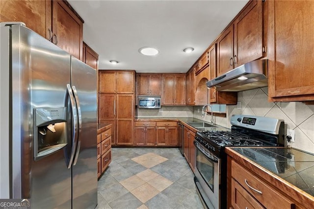 kitchen with decorative backsplash, appliances with stainless steel finishes, brown cabinets, under cabinet range hood, and a sink