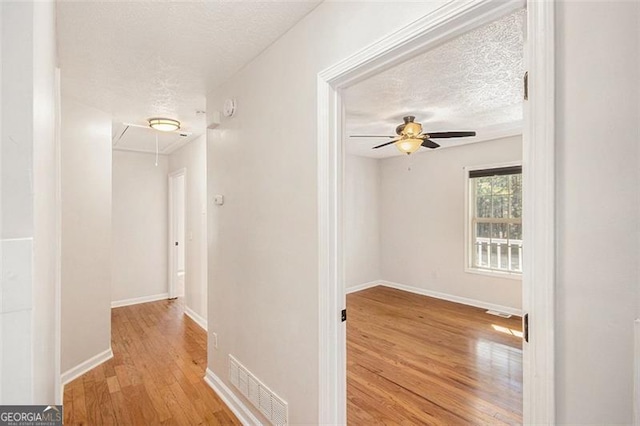 hall with a textured ceiling, wood finished floors, visible vents, baseboards, and attic access