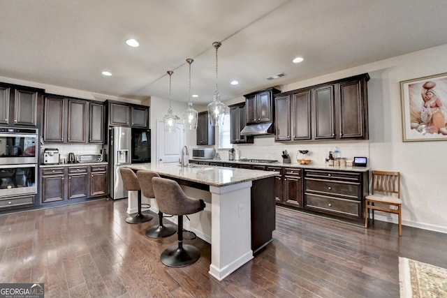 kitchen with appliances with stainless steel finishes, a breakfast bar, backsplash, under cabinet range hood, and dark wood-style floors