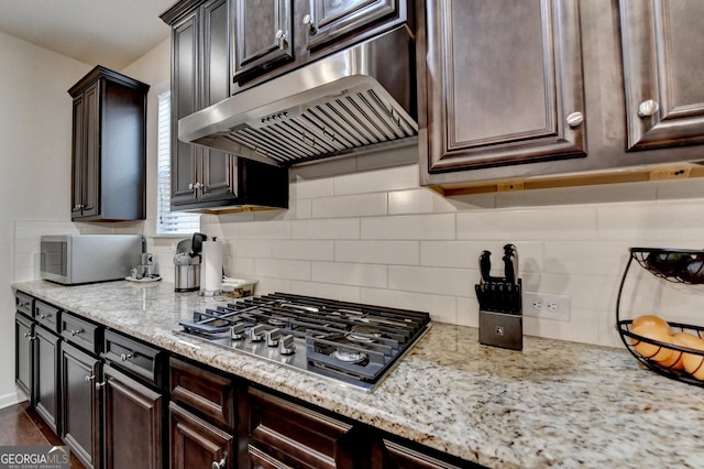 kitchen featuring decorative backsplash, dark brown cabinetry, appliances with stainless steel finishes, light stone countertops, and under cabinet range hood