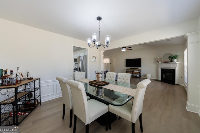 dining area featuring a decorative wall, ceiling fan with notable chandelier, wood finished floors, a fireplace with flush hearth, and ornate columns