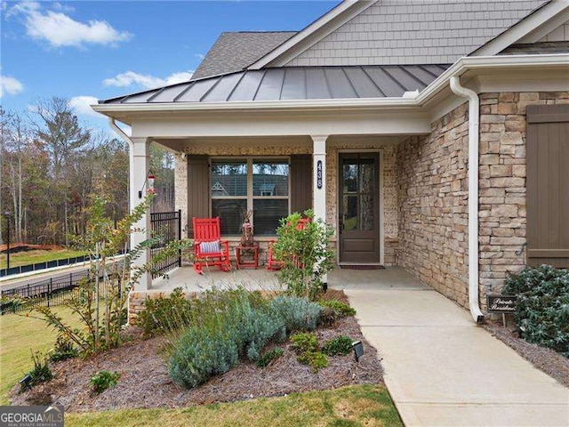 doorway to property with fence, a standing seam roof, a porch, stone siding, and metal roof